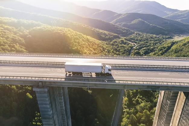 Caminhão branco cruzando uma ponte elevada cercada por montanhas e vegetação densa, representando logística eficiente em trajetos de longa distância.